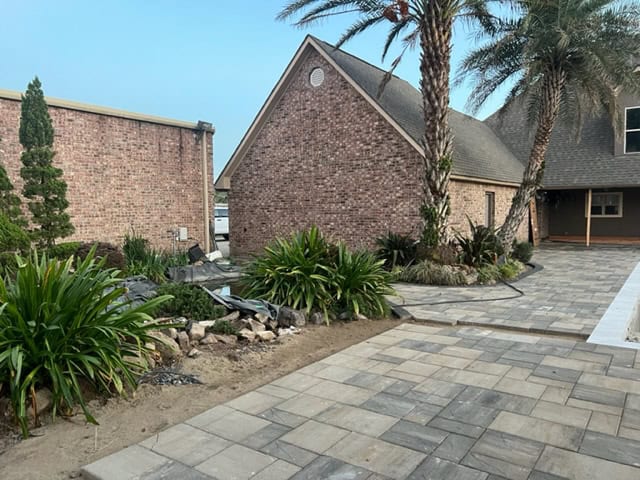 A neatly landscaped backyard with brick buildings, stone pathways, palm trees, and various plants. The area includes a small water feature on the left side, with rocks around it and well-maintained greenery. A spacious stone patio enhances the charm of this sunny oasis under a clear sky.
