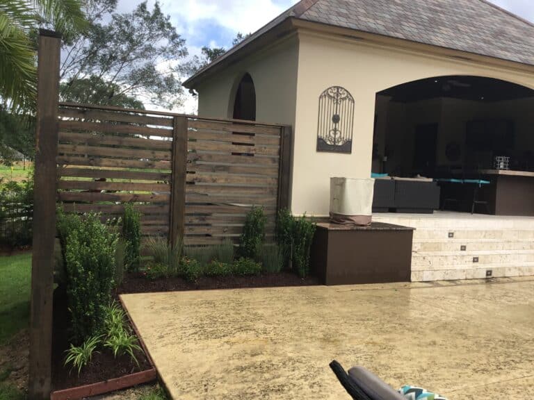 A backyard patio area featuring an outdoor seating space under a covered structure. The area is decorated with green plants, small shrubs, and a rustic wooden fence for added privacy. There is a staircase leading up to the seating area and a stained concrete floor in the foreground, offering screening from neighbors.