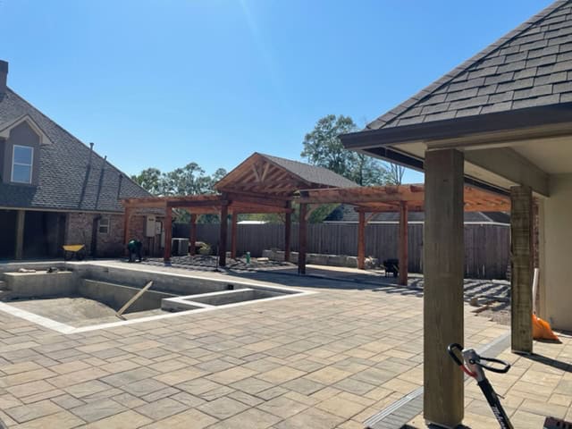 A large backyard under renovation features a partially built wooden pergola and an empty, rectangular in-ground pool. The yard is lined with stone tiles, forming a charming patio area. Houses and a fence stand in the background under a clear blue sky.
