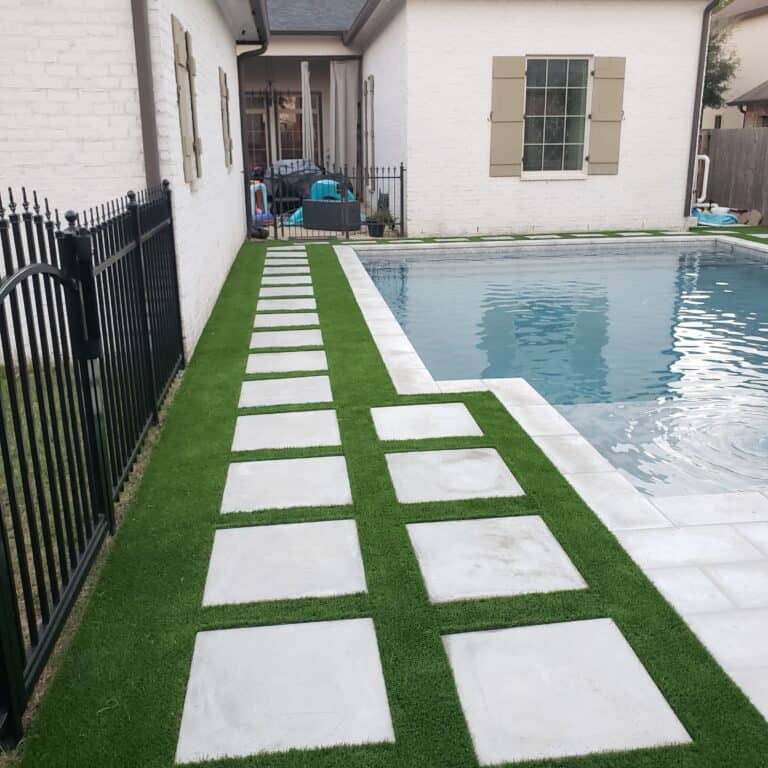 A backyard pool with clear water is adjacent to a house with a white brick exterior. Surrounding the pool are green strips of synthetic turf inlaid with large square concrete pavers forming a pathway. A black metal fence runs along the left side of the pool area.