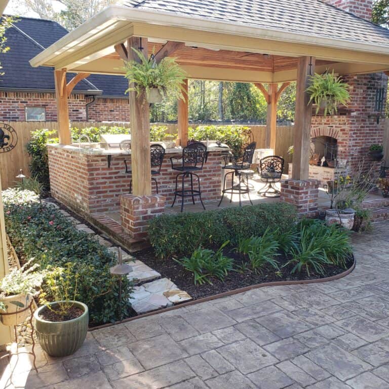 A backyard with a covered patio area featuring a brick structure, bar stools, and hanging plants. The patio has a tiled roof extension and is surrounded by a brick floor and garden beds with plants. This inviting space, reminiscent of charming pergolas, offers ample seating and a cozy atmosphere.