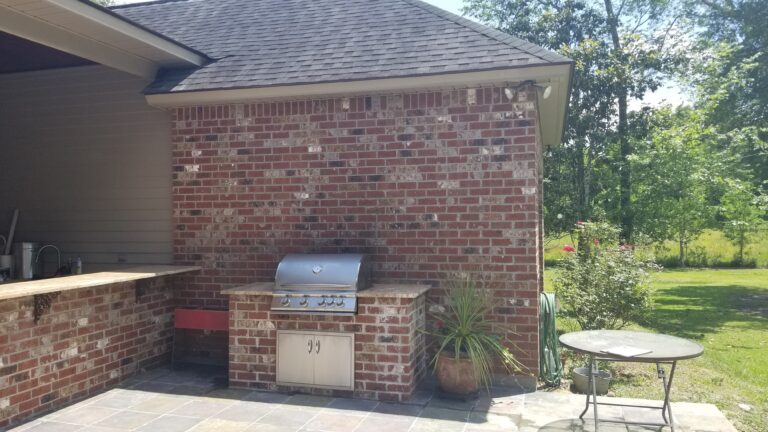 A backyard patio features an outdoor kitchen with a built-in stainless steel grill set into a brick structure, a small countertop to the left with a sink, a potted plant on the ground, a round patio table, and a garden hose nearby. Trees are in the background.