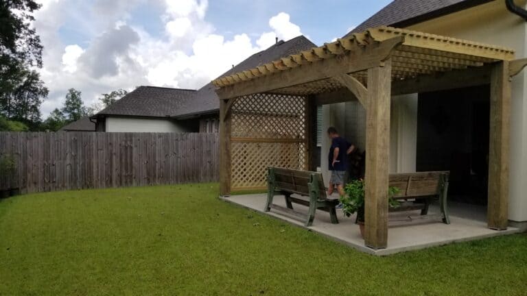 A backyard with a wooden pergola over a concrete patio. The pergola, featuring a lattice wall on one side, provides shade for a table with benches beneath it. A man stands near the structure, partially facing away. The yard is surrounded by a wooden fence, with houses visible in the background.