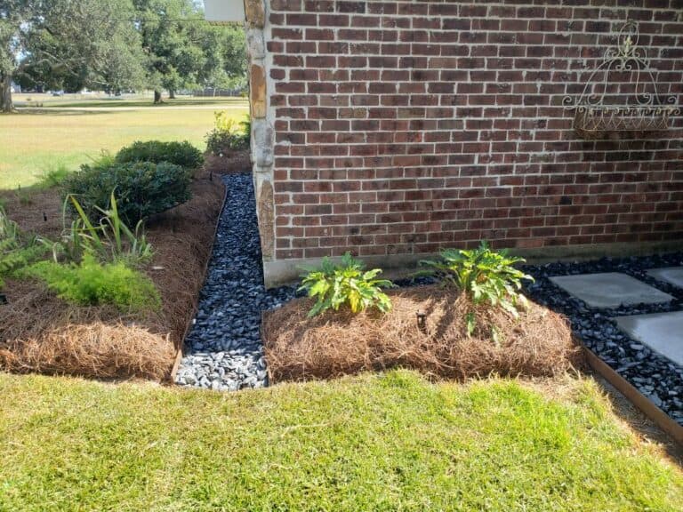 A landscaped garden adjacent to a brick wall features stone pathways and dry creek beds surrounded by mulch and greenery. Bushes and small plants are planted in the mulch beds, and the lawn is well-maintained. The area is bordered by trees in the background.
