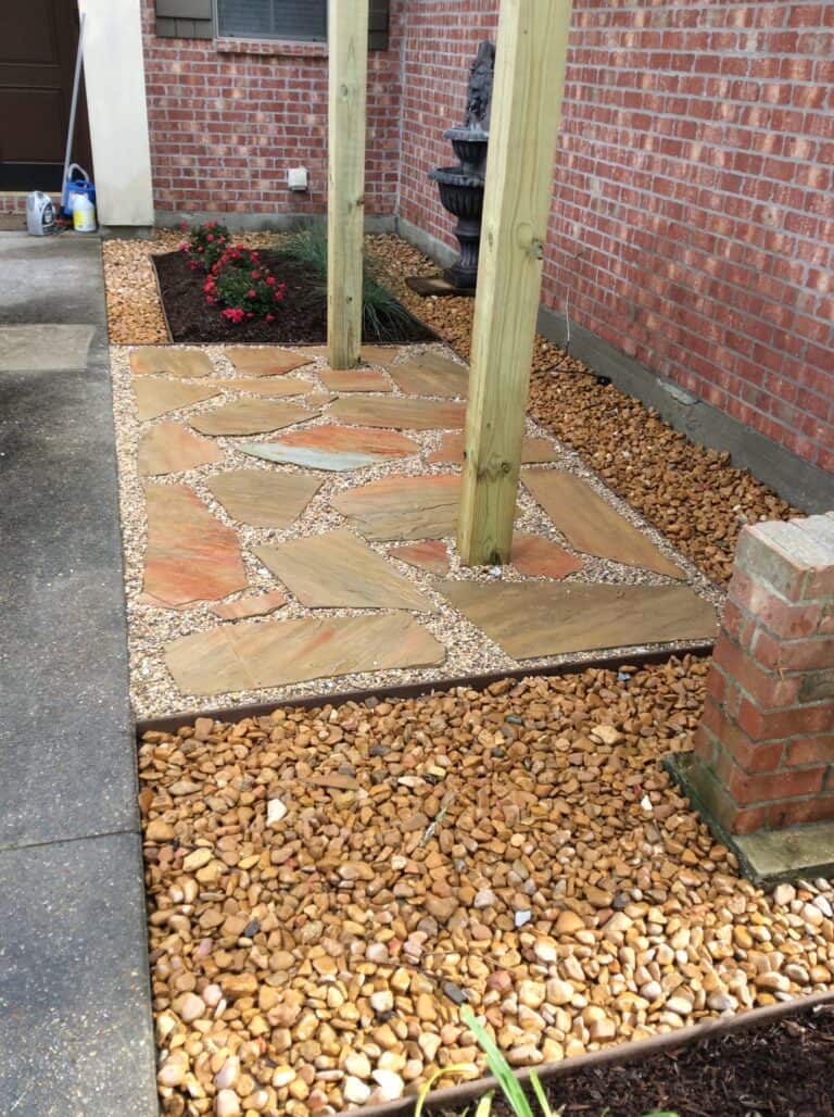 A garden path made of irregularly shaped natural stone slabs is surrounded by small pebbles and larger river rocks. Wooden posts support an overhead structure. Red brick walls and flowerbeds with red flowers border the path.