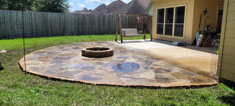 A fabulous fire pit sits on a curved flagstone surface in the backyard patio. Extending from a house with yellow siding, the patio is adjacent to a concrete area with a wooden swing. Enclosed by a wooden fence and featuring well-maintained grass, this outdoor space offers perfect outdoor heating.