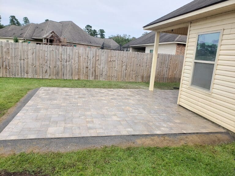 A spacious backyard patio with light-colored stone pavers sits next to a beige house with a double-door entrance. The patio, bordered by a grassy lawn and enclosed by a wooden privacy fence, offers glimpses of neighboring houses in the background.