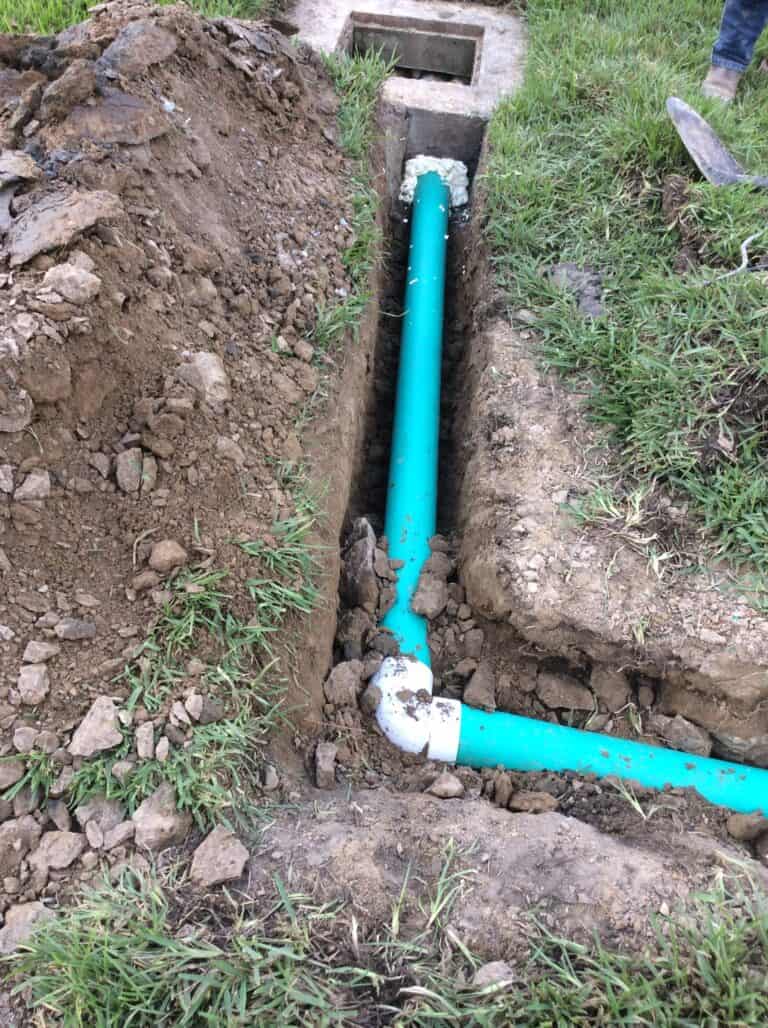 A dug trench containing a newly installed blue PVC water pipe with a white elbow joint, partially surrounded by soil and patches of grass. The subsurface trench appears to be in a residential yard, and a small concrete section is visible at the top.