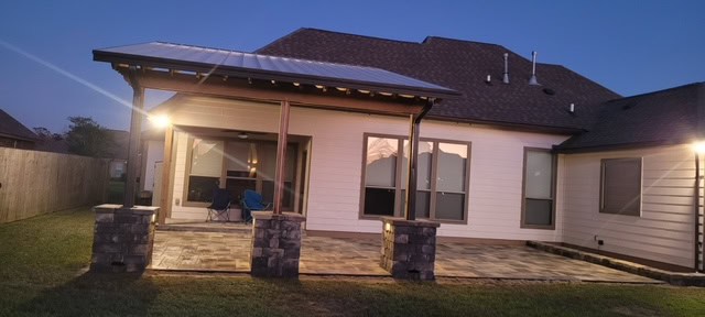 A backyard view at dusk showcasing a stone patio area attached to a house with white siding and a steep brown roof. The patio has stone pillars and outdoor seating. The area is illuminated by two exterior lights creating a warm ambiance.