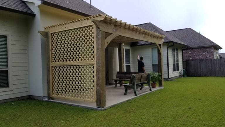 A wooden pergola with a lattice panel on one side is attached to the back of a house. There are two wooden benches beneath the pergola, and a person facing away from the camera stands next to one bench, pointing at something on the house wall, perhaps considering future roof extensions.