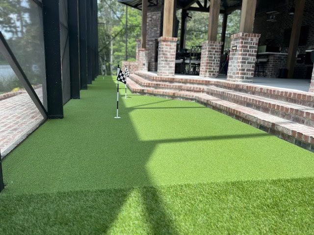 The image shows an inviting outdoor putting green next to a charming patio area with brick columns and stairs. The putting green, equipped with two holes marked by small checkered flags, is enclosed within a screened structure and surrounded by trees, creating an idyllic setting for practicing your putting skills.