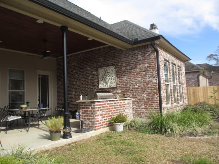 A brick house with a covered patio features an outdoor kitchen complete with a grill and table with chairs. The grill is built into a brick counter adorned with decorative artwork above it. Potted plants decorate the patio, which overlooks a grassy yard enclosed by a wooden fence. The sky is partly cloudy.