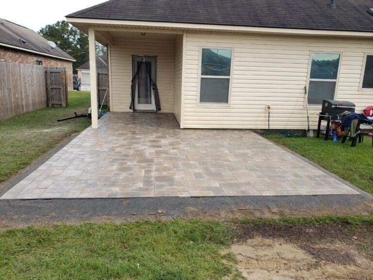 A stone patio with newly installed beige and grey pavers extends from a covered porch attached to a house. The backyard area is bordered by grass and fenced in, with some outdoor furniture and equipment visible on the right side near the house.