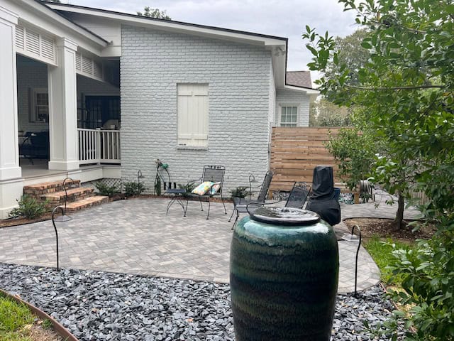 A backyard patio with gray pavers features black wrought iron chairs and a fire pit. In the foreground, a large green ceramic water feature stands beside a bed of dark gray gravel. For added privacy, trees and plants provide natural screening around the area, complementing the house’s covered porch and gray brick exterior.