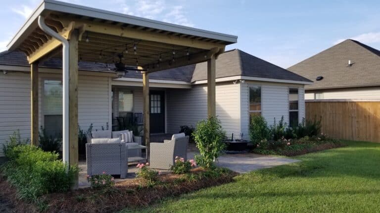 Backyard with a covered patio, pergolas, seating area, and garden. The house features light-colored siding, shingled roof, and a wooden fence.