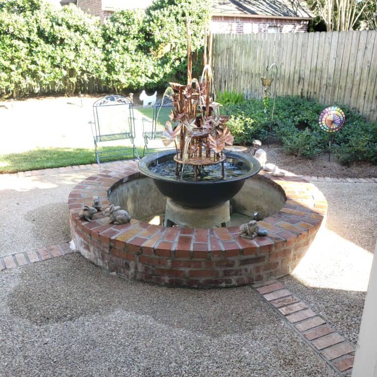A garden with a circular brick water fountain centerpiece. The fountain, one of the stunning water features, contains dried plants, and there are small statues of animals on the fountain edge. Metal chairs and garden decor, including a colorful spinning wheel, are arranged around the area.