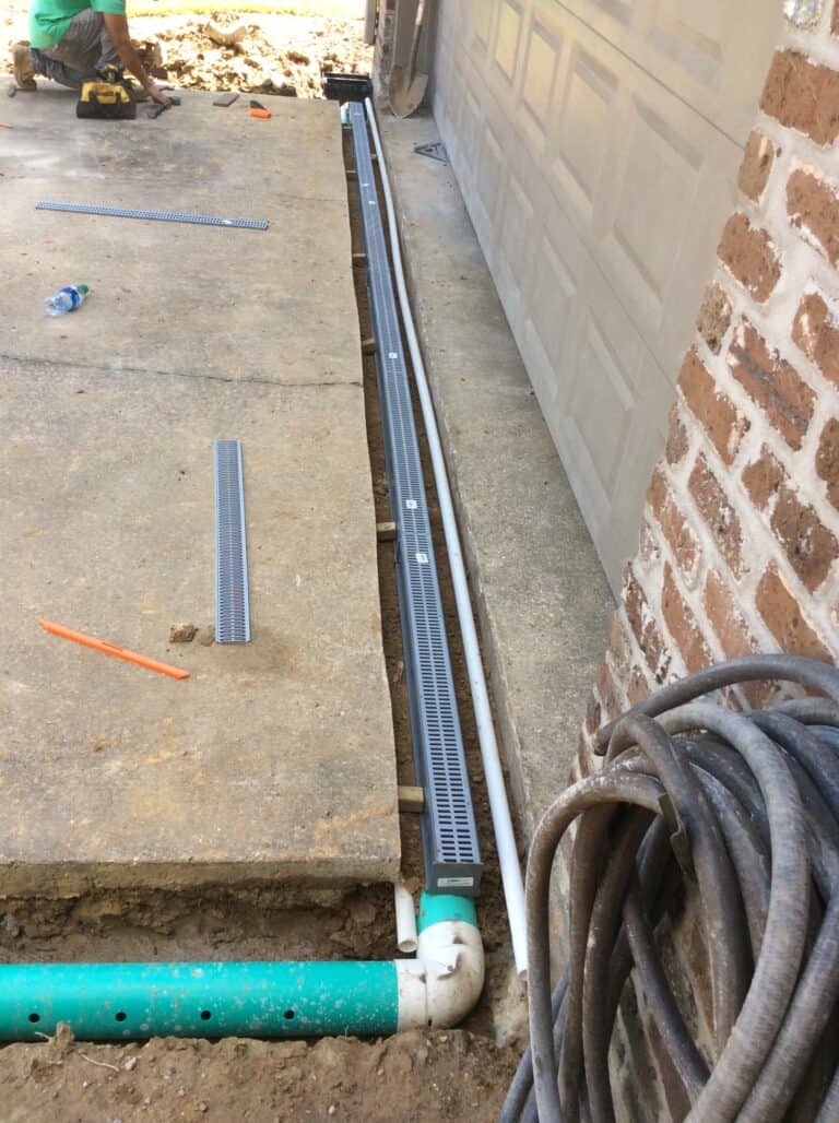 A construction site with a custom drainage system being installed beside a brick wall. A trench with green pipes is visible, and a gray drainage grate is being positioned into the subsurface ground. A worker in a green shirt is kneeling in the background, working with tools.