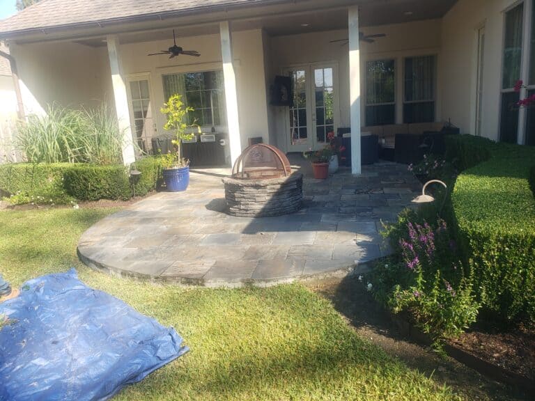 A fabulous backyard patio featuring a round stone fire pit and a mix of potted plants. The patio has stone flooring and is covered by a roof. There is a small, well-maintained lawn surrounding the patio area, with a blue tarp spread on the grass to the left.
