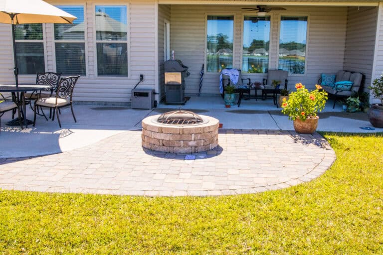 A fabulous backyard patio featuring a brick fire pit surrounded by a paved seating area. On the left, there is a table with chairs and an umbrella. The right side has a cozy seating area with a couch, chairs, and potted plants. The patio is adjacent to a house with large windows.
