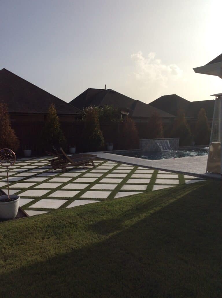 A backyard pool area with a geometric-patterned patio featuring two reclined sun loungers. The pool has a waterfall feature, bordered by lush synthetic turf. The sky appears overcast, and there are several houses and trees in the background.