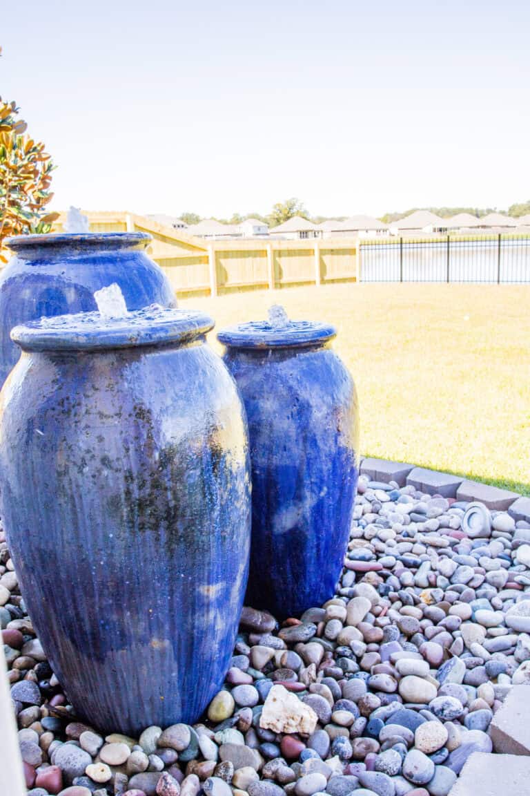 Three large blue ceramic jars with water fountains on top stand as stunning water features on a bed of smooth, multicolored pebbles in an outdoor space. A wooden fence and grassy yard can be seen in the background under a clear sky.