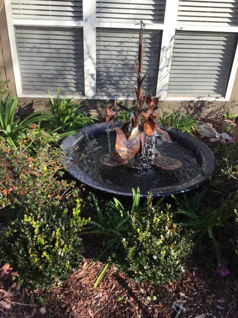 A small outdoor fountain with metal leaf sculptures stands in the center of a circular basin filled with water, surrounded by green plants and shrubs. This enchanting water feature is in front of a house with white window blinds partially visible in the background.