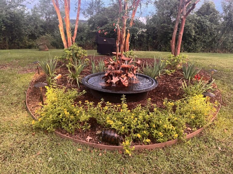 A garden fountain, a stunning water feature, is surrounded by flowers and mulch in a circular arrangement, with trees and grass in the background.