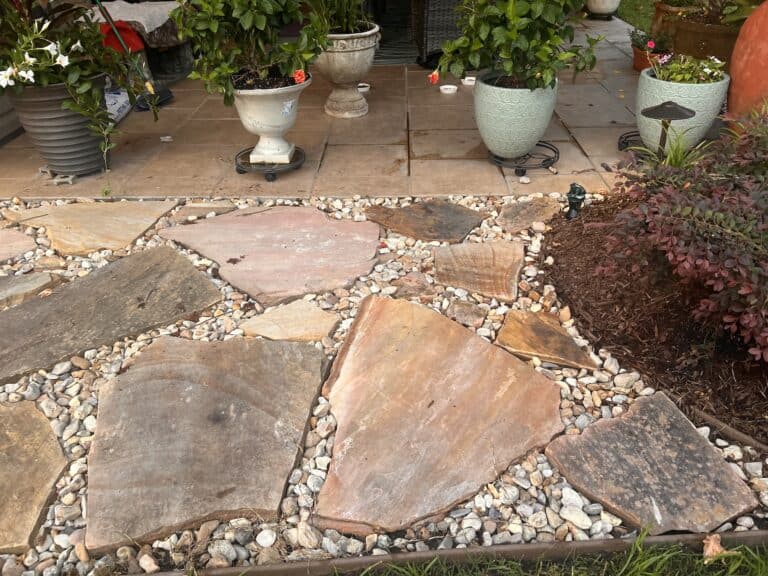 Natural stone path with large, irregular stones surrounded by small pebbles. Lush plants in pots and a small garden bed enrich the background.
