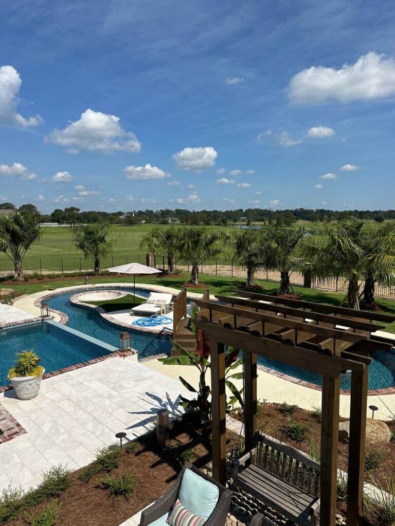A backyard view with a swimming pool, palm trees, and a pergola housing a cozy daybed swing overlooks a grassy field beyond the fence under a partly cloudy sky.