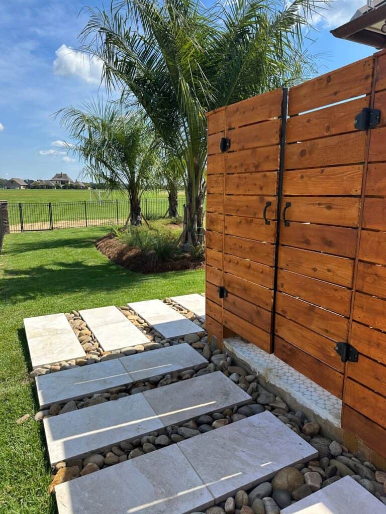 A pathway of rectangular stone slabs, surrounded by pebbles, a wooden fence for privacy screening, and palm trees in a grassy yard.
