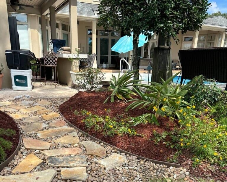 A backyard oasis with a stone pathway winds through lush plants and red mulch, leading to a serene pool area. Patio furniture sits invitingly under an umbrella, while dry creek landscaping adds rustic charm to the scene.