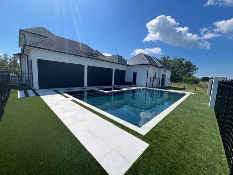 A modern house with a dark roof and a fenced backyard showcases a rectangular swimming pool surrounded by synthetic turf and concrete tiles.