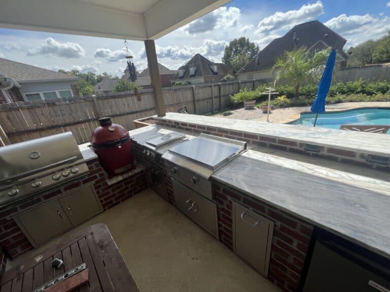 This stunning outdoor kitchen features a grill, smoker, prep area, and sink on a stone countertop, all overlooking a backyard with a pool and elegant patio furniture.