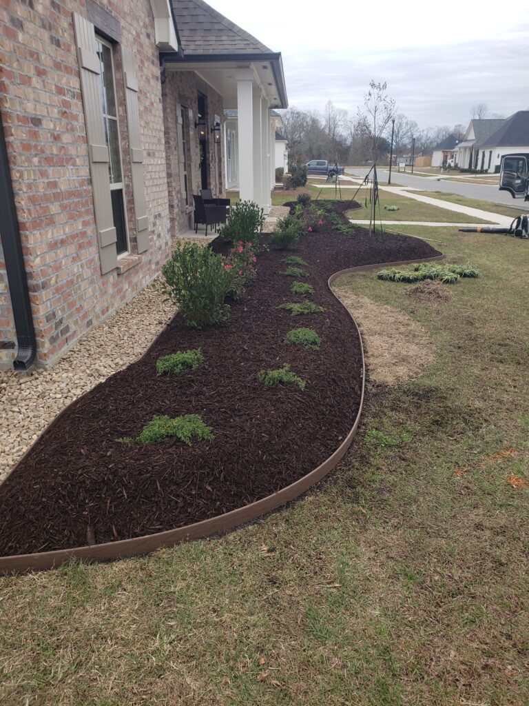 A freshly landscaped garden showcases a mulched flower bed with small, young plants neatly lined by landscape borders in front of a brick house with windows and shutters. A driveway and other houses can be seen in the background under a cloudy sky.
