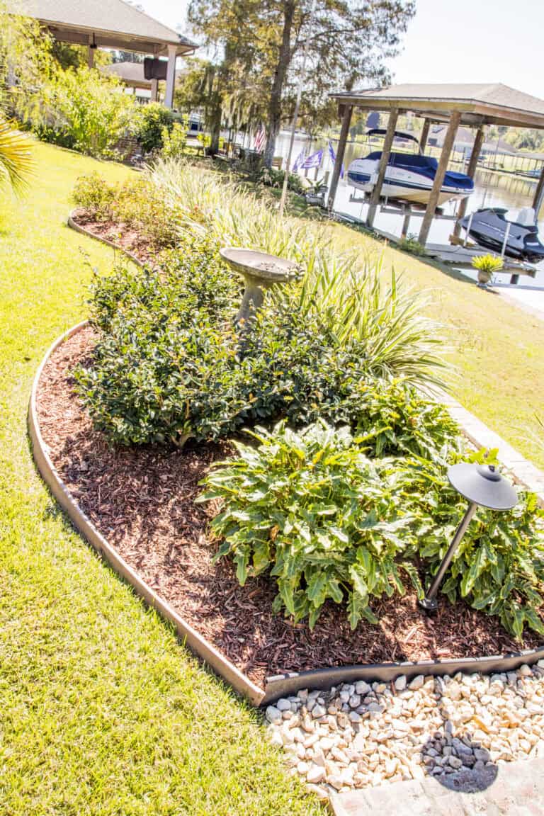 A well-manicured garden with a birdbath at the center, surrounded by various shrubs and plants, mulched with wood chips. Landscape borders enhance the stone pathway leading to a waterside dock with boats, partially shaded by trees and a pergola in the background.