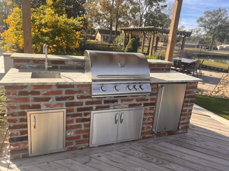 A modern outdoor kitchen featuring a built-in stainless steel gas grill, a sink with a faucet, and storage cabinets below. The setup is made of red brick and sits on a wooden deck with a garden and pergola visible in the background.