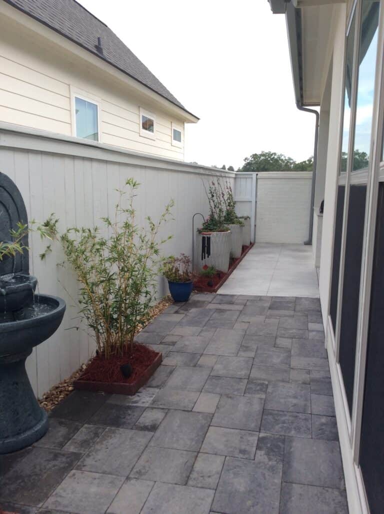 Narrow backyard space with decorative stone tiles, potted plants along a white fence, a small fountain, and adjacent house walls.