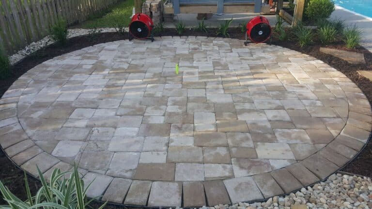 Circular hardscape patio with stone tiles, bordered by small rocks and plants. Two red air movers are positioned opposite each other on top.