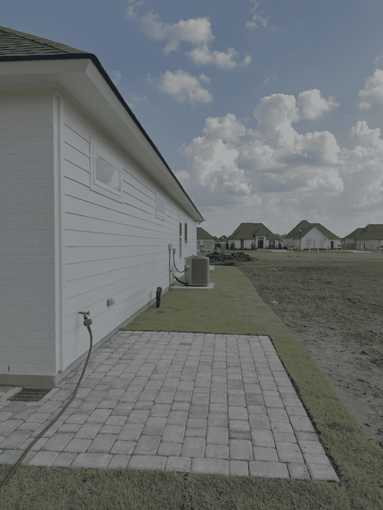 Side view of a white house with a brick hardscape patio, hose, air conditioning unit, and open grassy backyard; several homes in the background.