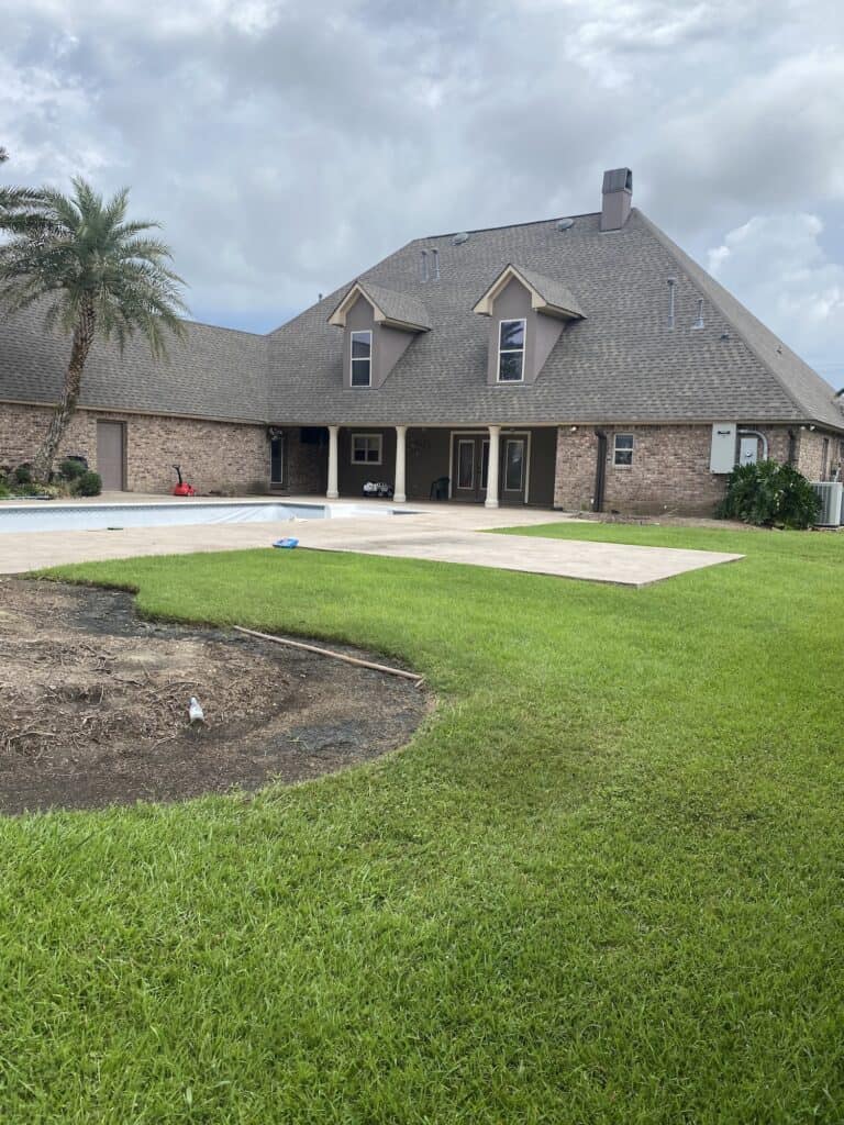 A large brick house with a steep roof, covered patio, swimming pool, and a well-maintained lawn under a cloudy sky.