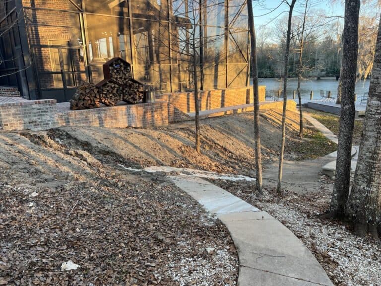 Curved concrete walkway beside a brick building with stacked firewood, trees, and a river in the background.