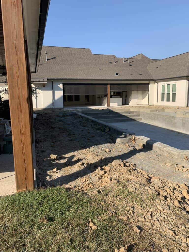 A partially built backyard pool surrounded by dirt and construction materials near a large house with a covered patio.