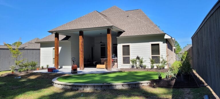 A backyard with a covered patio, artificial turf putting green, plants, and a wooden fence under a clear blue sky.