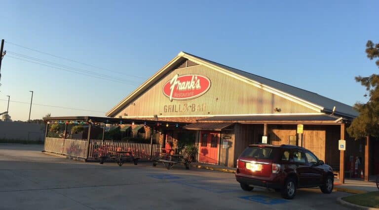 A red SUV is parked in front of Frank's Grill & Bar, a rustic, metal-roofed restaurant with a covered outdoor seating area.