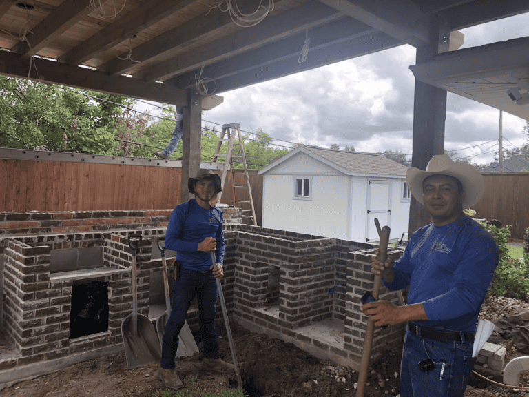 Two men in blue shirts and hats work with shovels at a Crescent Courtyard brick outdoor kitchen under a wooden patio cover.