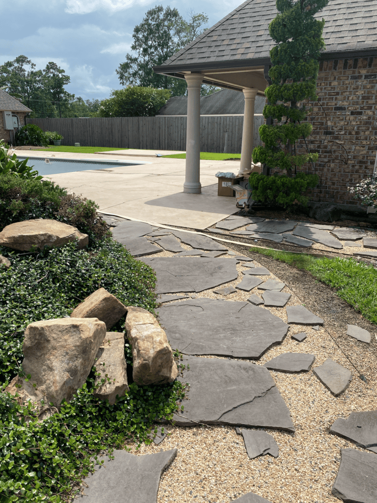 A stone path leads past landscaping and rocks toward a covered patio near a pool in a suburban backyard.
