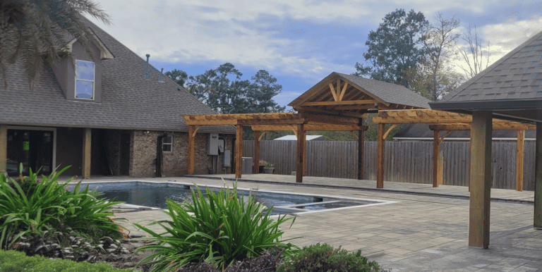 Outdoor pool area at Pavillion Pointe with a wooden pergola, paved patio, lush plants, and two sloped-roof buildings under a partly cloudy sky.