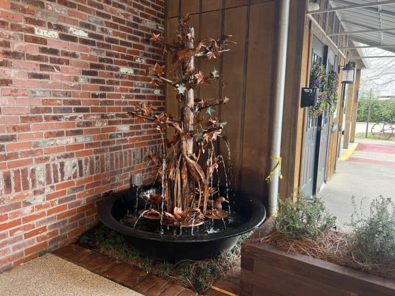 A metal tree fountain with water streams stands in a black basin near a brick and wood wall outside a building entrance.