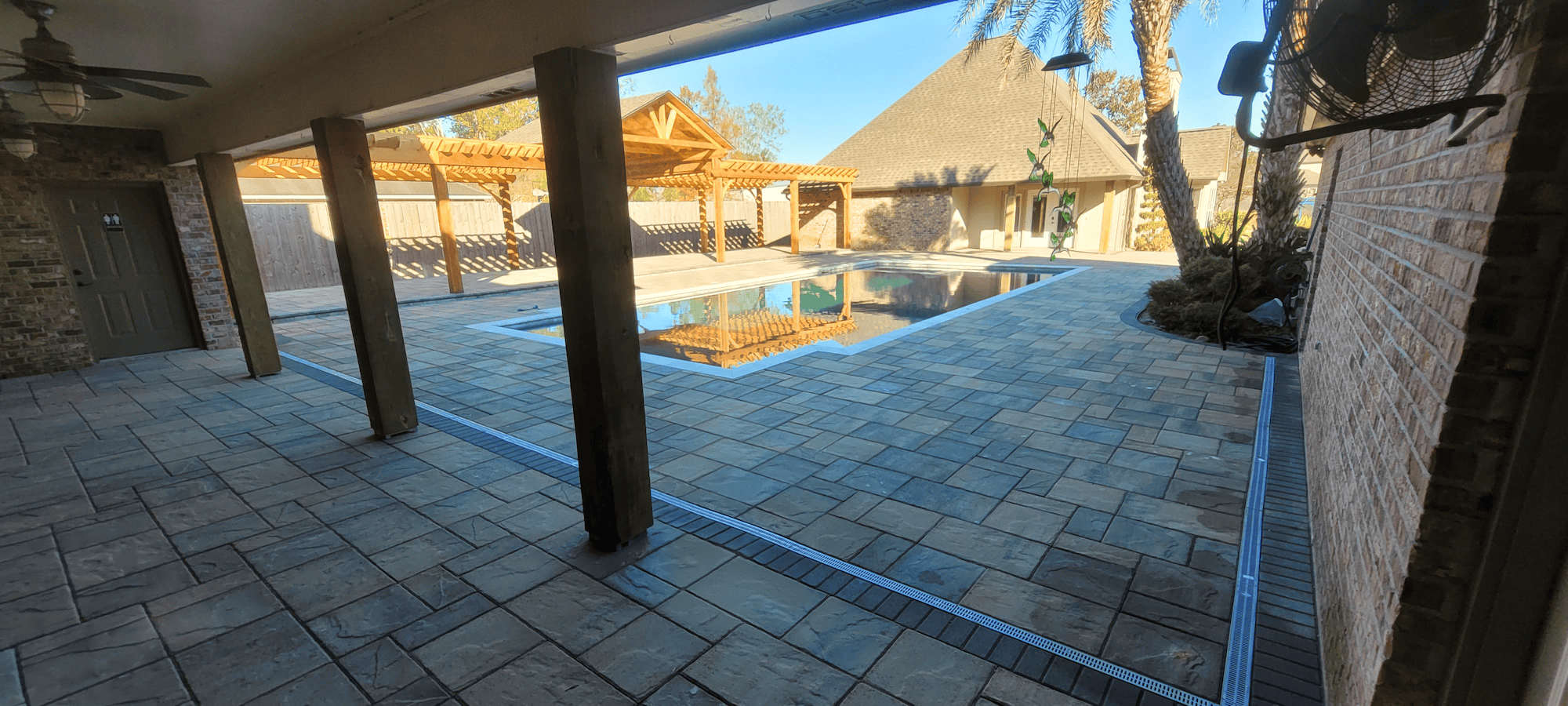 Covered patio with stone tile floor overlooking a pool, pergola, and outbuilding in a fenced backyard with palm trees.