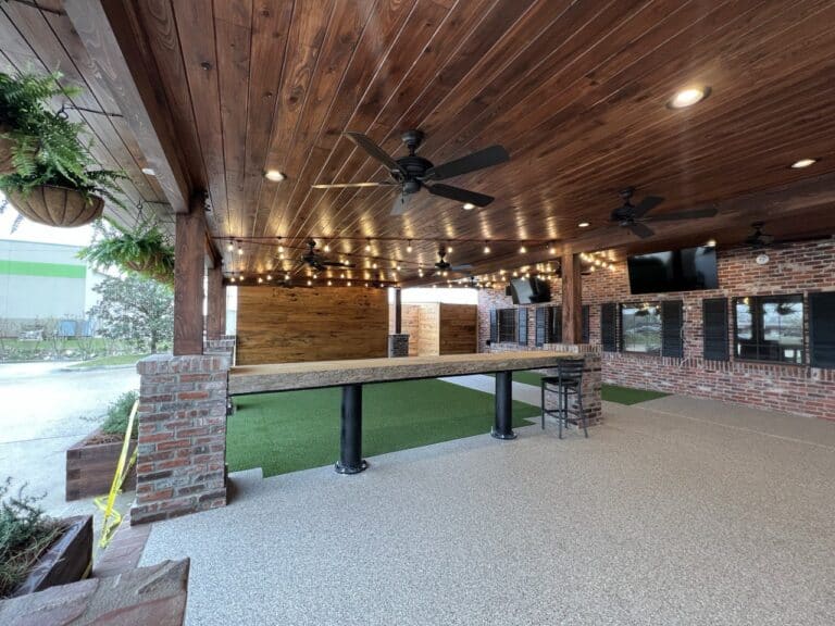 Covered outdoor patio with wooden ceiling, string lights, ceiling fans, brick walls, artificial turf, and bar-height tables and stools.